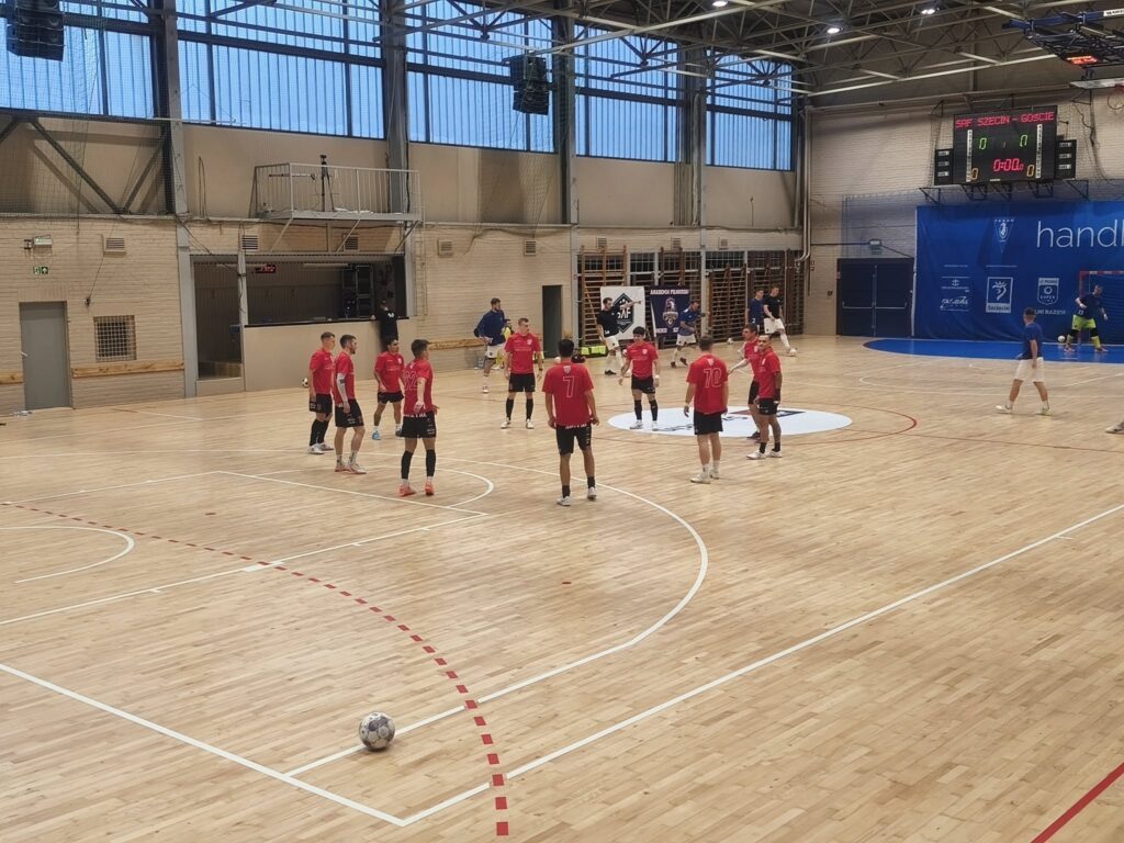 Zawodnicy w czerwonych strojach trenują na boisku futsalowym W tle widoczna tablica wyników.