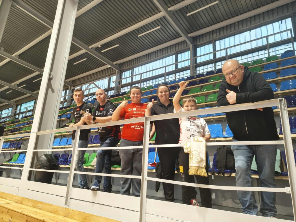 Grupa kibiców Futsal Leszno w hali sportowej, uśmiechają się i podnoszą kciuki w górę, na tle kolorowych siedzeń.